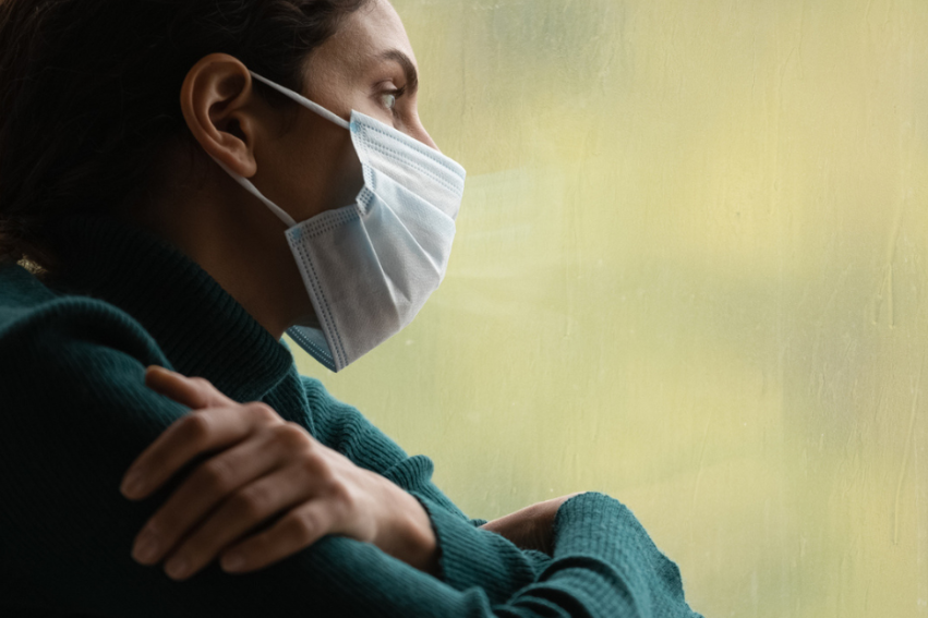 Woman in a mask looks out a window pensively. She wears a green sweater and rests her arms on each other