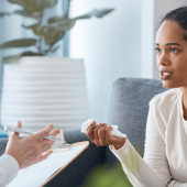 A woman in a white shirt sits on a sofa, intently talking to a therapist holding a clipboard.