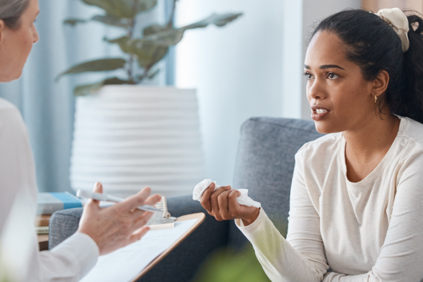 A woman in a white shirt sits on a sofa, intently talking to a therapist holding a clipboard.
