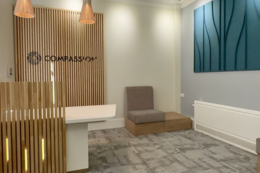 Modern waiting room with soft lighting and neutral tones. Features a wooden slat accent wall, minimalist seating, and abstract blue wall art