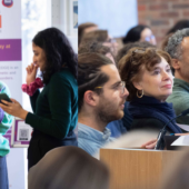 A diverse group gathers at an EDGI event. On the left, two smiling women in branded shirts engage with a smartphone. On the right, attendees listen attentively as a woman speaks at a podium.