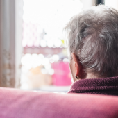 An elderly person with short gray hair sits in a cozy room, gazing out of a sunlit window