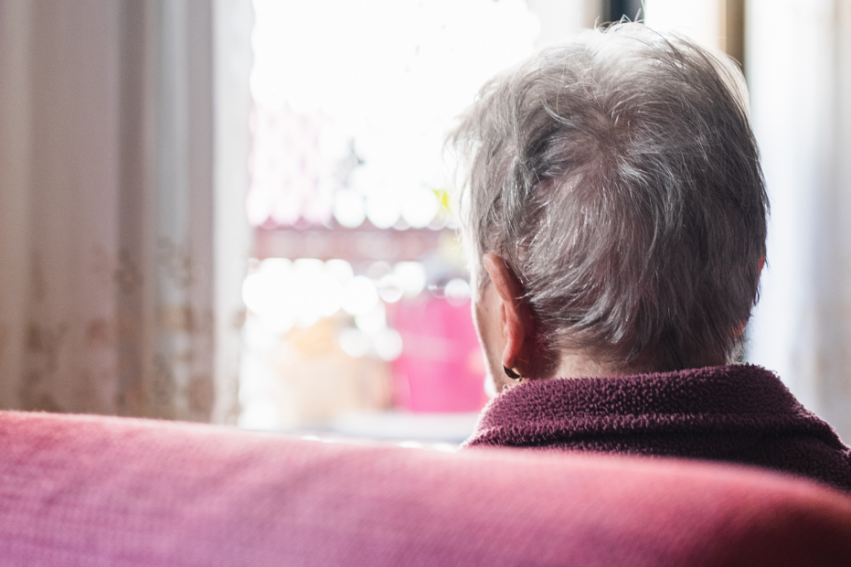 An elderly person with short gray hair sits in a cozy room, gazing out of a sunlit window