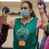 A caregiver, wearing a green apron and mask, assists two elderly women, also masked, in a joyous arm exercise. They are wearing face masks