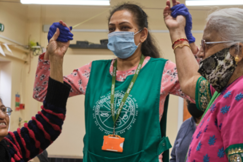 A caregiver, wearing a green apron and mask, assists two elderly women, also masked, in a joyous arm exercise. They are wearing face masks