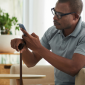 A man in a gray polo shirt sits on a sofa, using a cane and a smartphone