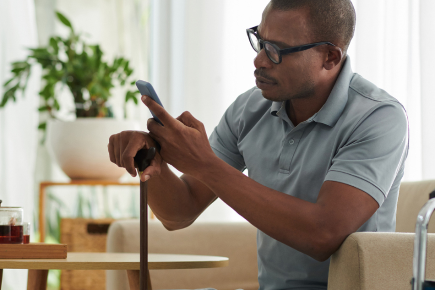 A man in a gray polo shirt sits on a sofa, using a cane and a smartphone