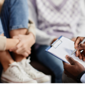 A therapist holds a clipboard and pen, engaging with two people seated closely, one with arms wrapped around their knees.
