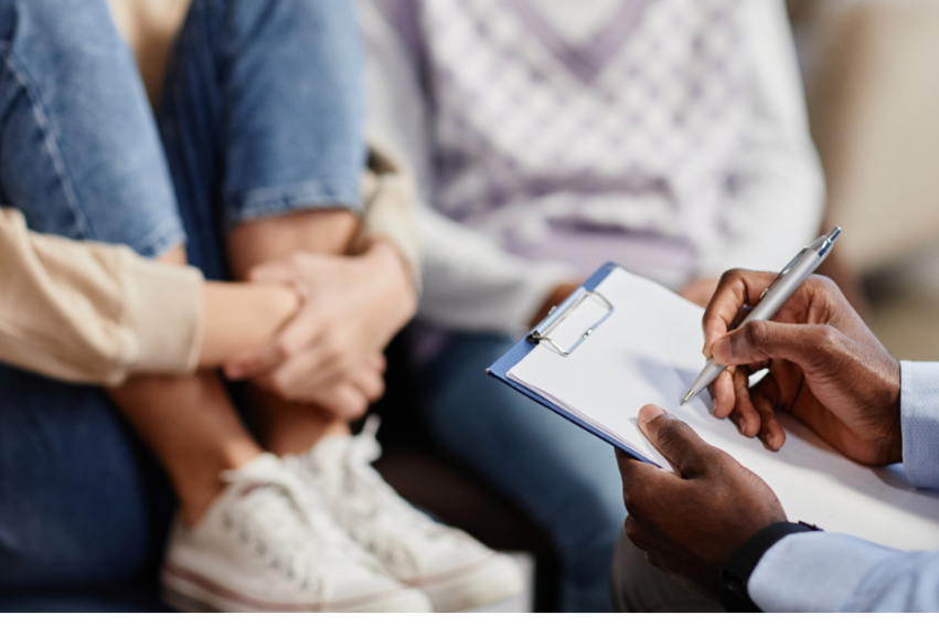 A therapist holds a clipboard and pen, engaging with two people seated closely, one with arms wrapped around their knees.