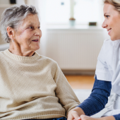 An elderly woman in a beige sweater smiles at a young female nurse in a white uniform