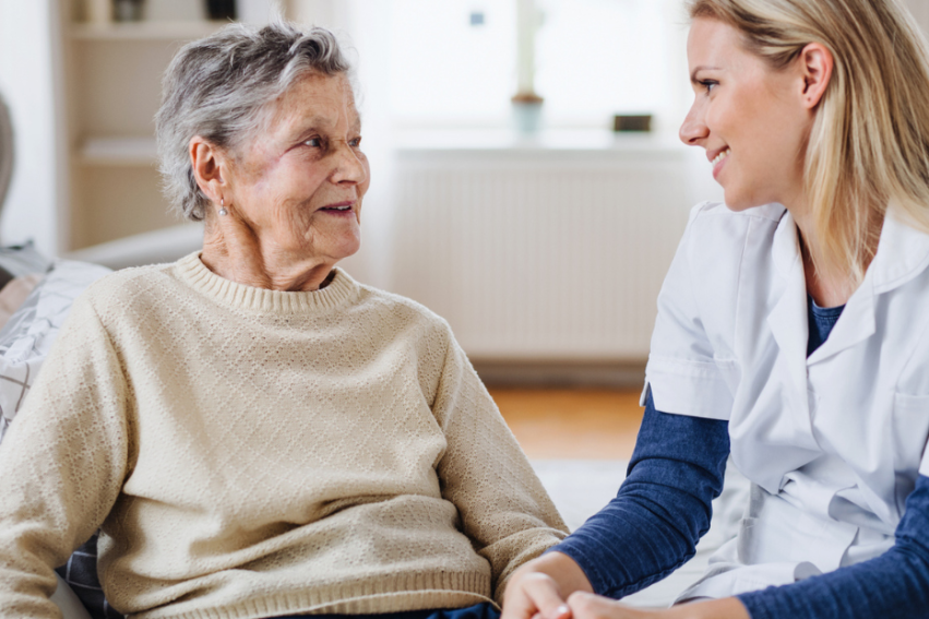 An elderly woman in a beige sweater smiles at a young female nurse in a white uniform