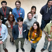 A diverse group of 18 people stands in a circle, smiling up at the camera