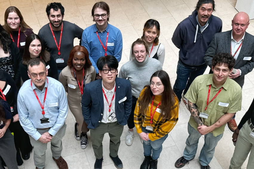A diverse group of 18 people stands in a circle, smiling up at the camera