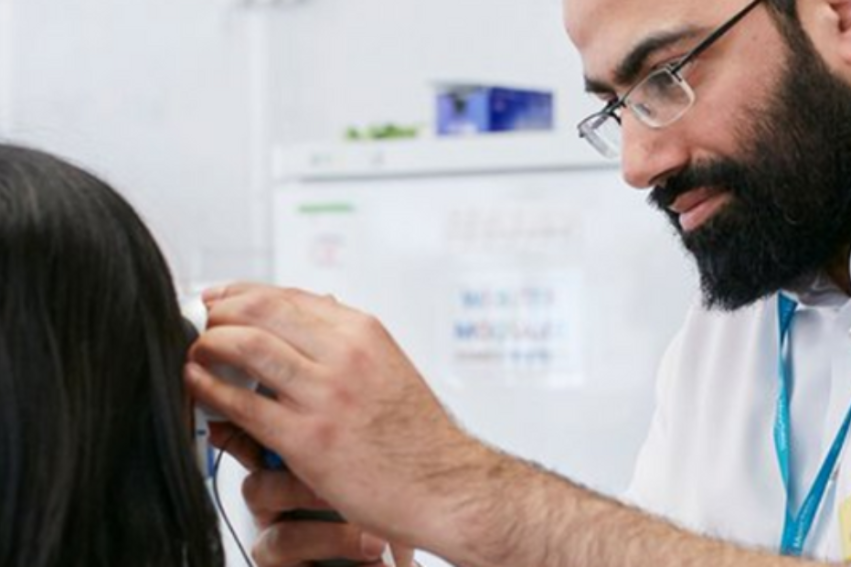 A healthcare professional with glasses and a beard examines a patient's ear in a clinical setting