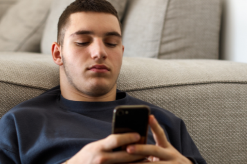 Young man lying on a gray couch scrolling on his smartphone