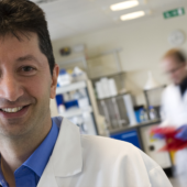 Smiling man in a white lab coat stands in a bright laboratory. Shelves and scientific equipment fill the room