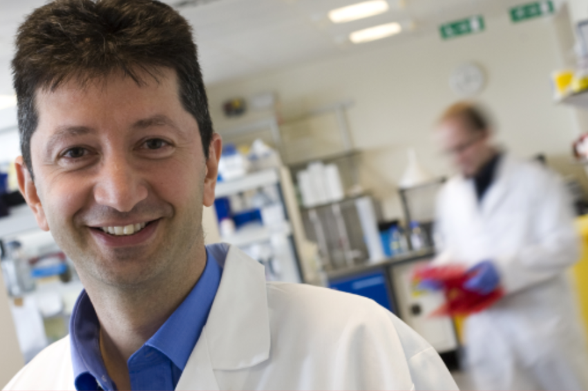 Smiling man in a white lab coat stands in a bright laboratory. Shelves and scientific equipment fill the room