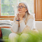 Elderly woman with gray hair and glasses looks thoughtful, sitting on a cozy sofa with colorful cushions