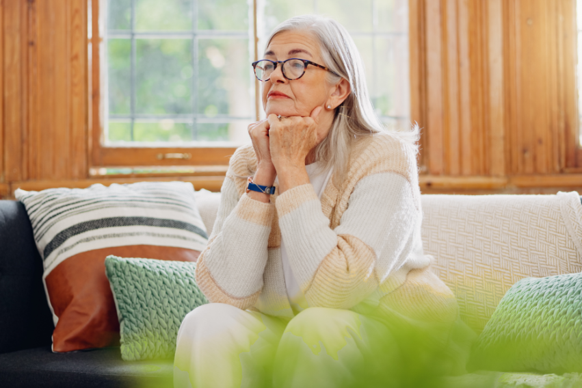 Elderly woman with gray hair and glasses looks thoughtful, sitting on a cozy sofa with colorful cushions