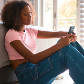 A young woman in a pink shirt and jeans sits on the floor, leaning against a bed, using her phone.