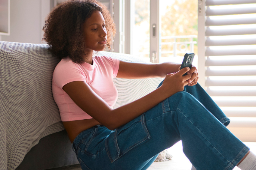A young woman in a pink shirt and jeans sits on the floor, leaning against a bed, using her phone.