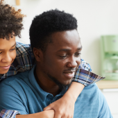 Father and son smiling, with the son playfully hugging the father from behind