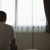 A man sits on a bed, facing a window with sheer curtains, in a dimly lit room