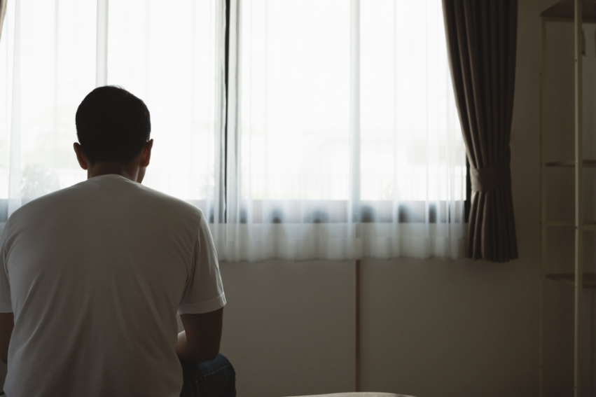 A man sits on a bed, facing a window with sheer curtains, in a dimly lit room