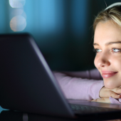 A smiling young girl with headphones watches a laptop in a dimly lit room