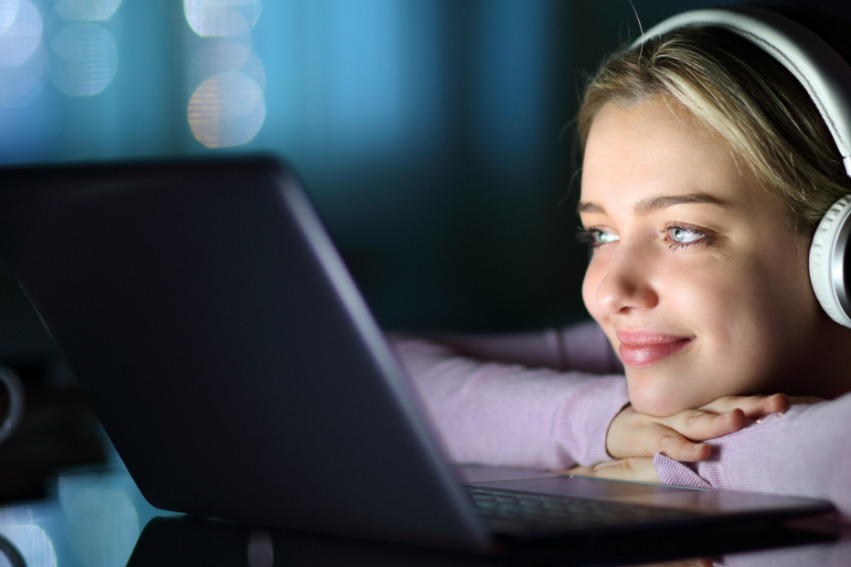 A smiling young girl with headphones watches a laptop in a dimly lit room