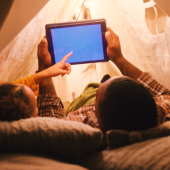 Cozy indoor tent with soft lighting; father and a daughter inside, lying side-by-side, looking at a tablet.