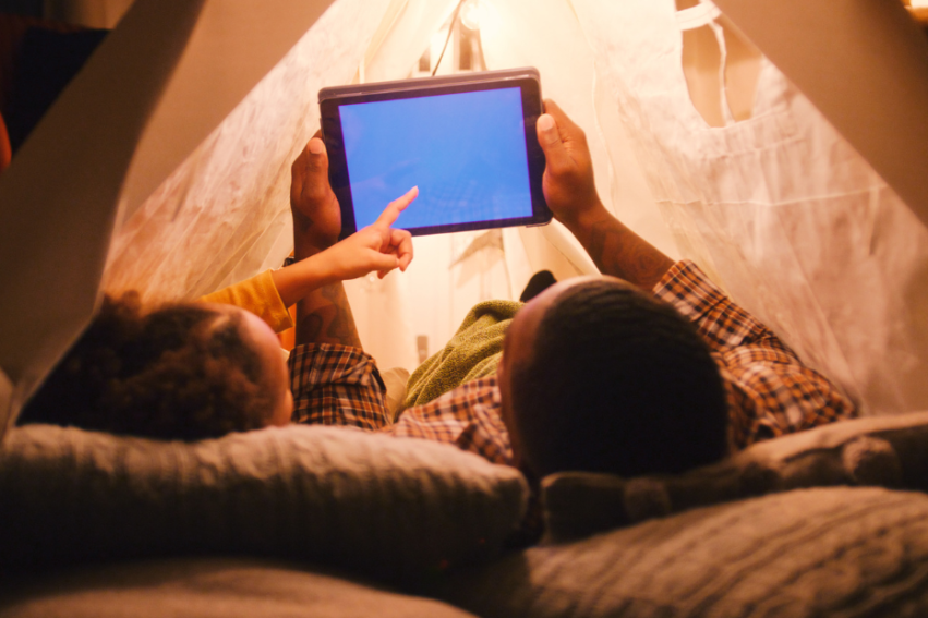 Cozy indoor tent with soft lighting; father and a daughter inside, lying side-by-side, looking at a tablet.