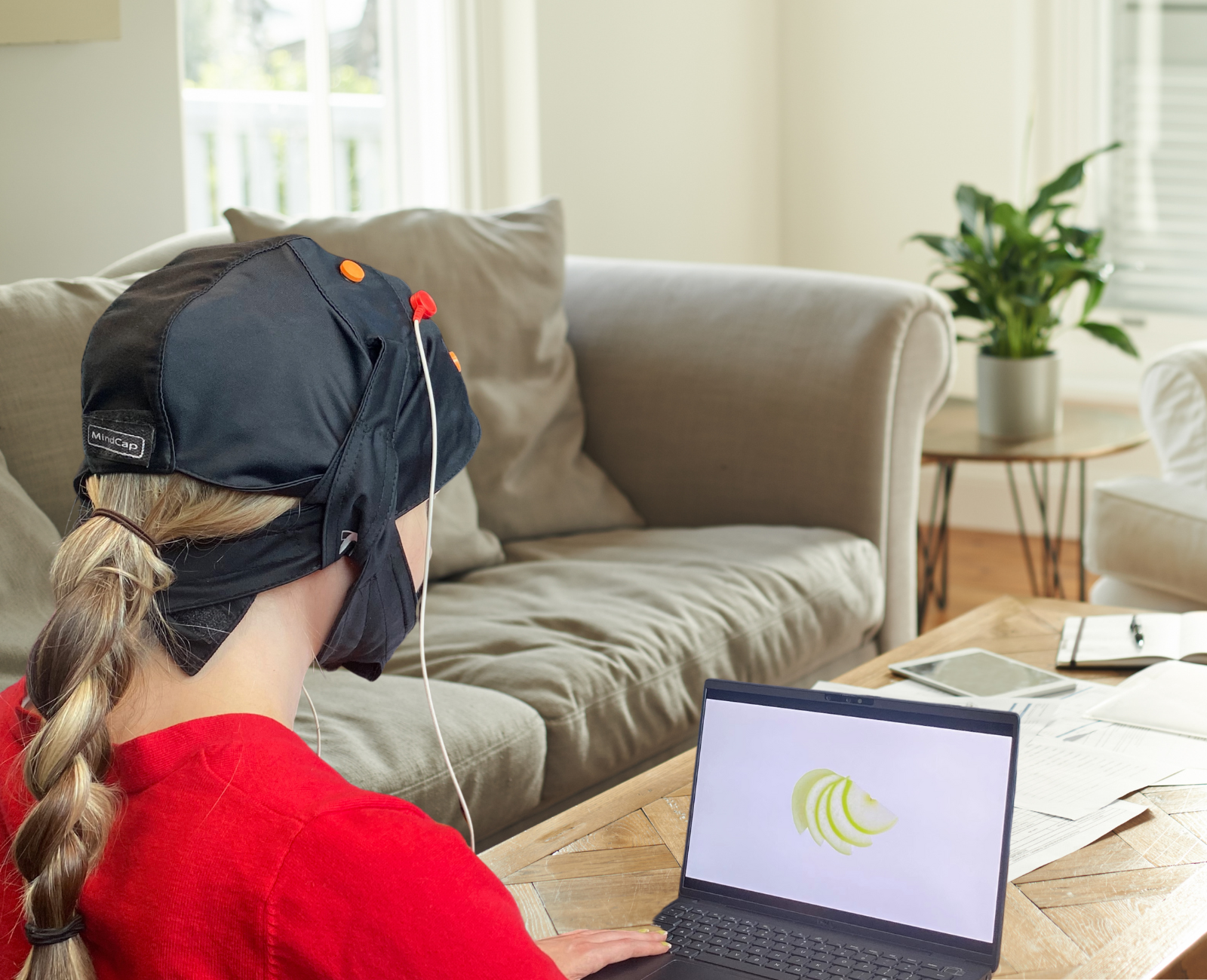 a woman sits at home looking at a laptop. she is wearing a black cloth cap strapped under her chin, with a wire coming off it