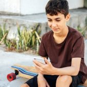 Teen boy sitting on skateboard using his smartphone