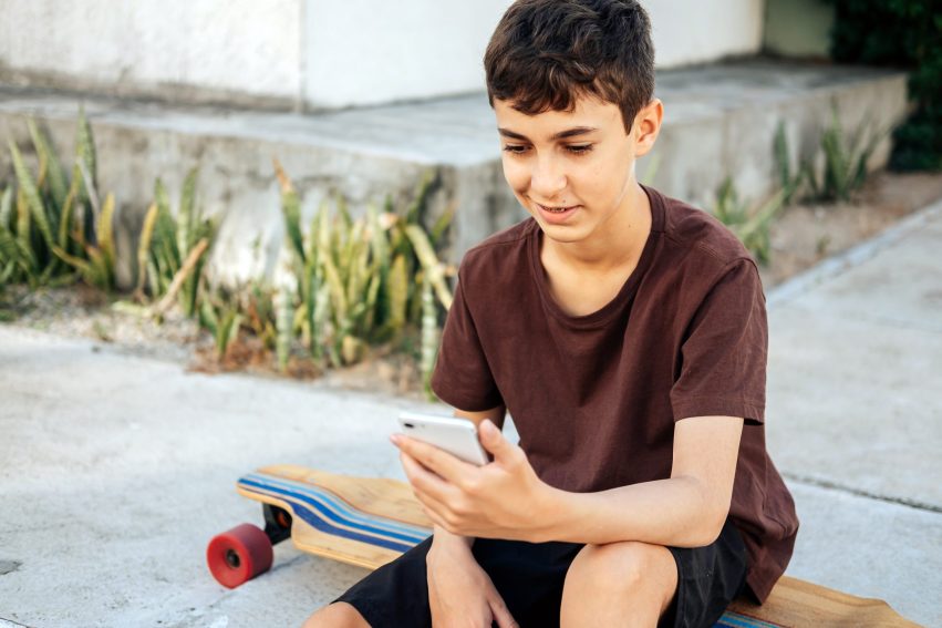 Teen boy sitting on skateboard using his smartphone