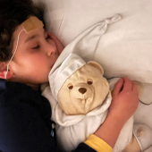 A child sleeps on a white pillow, hugging a teddy bear while connected to medical sensors
