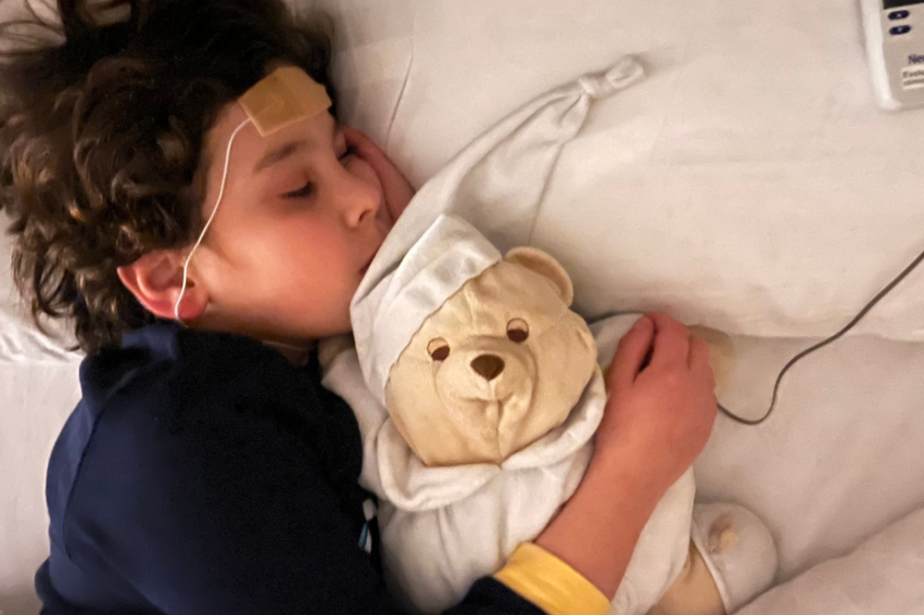 A child sleeps on a white pillow, hugging a teddy bear while connected to medical sensors